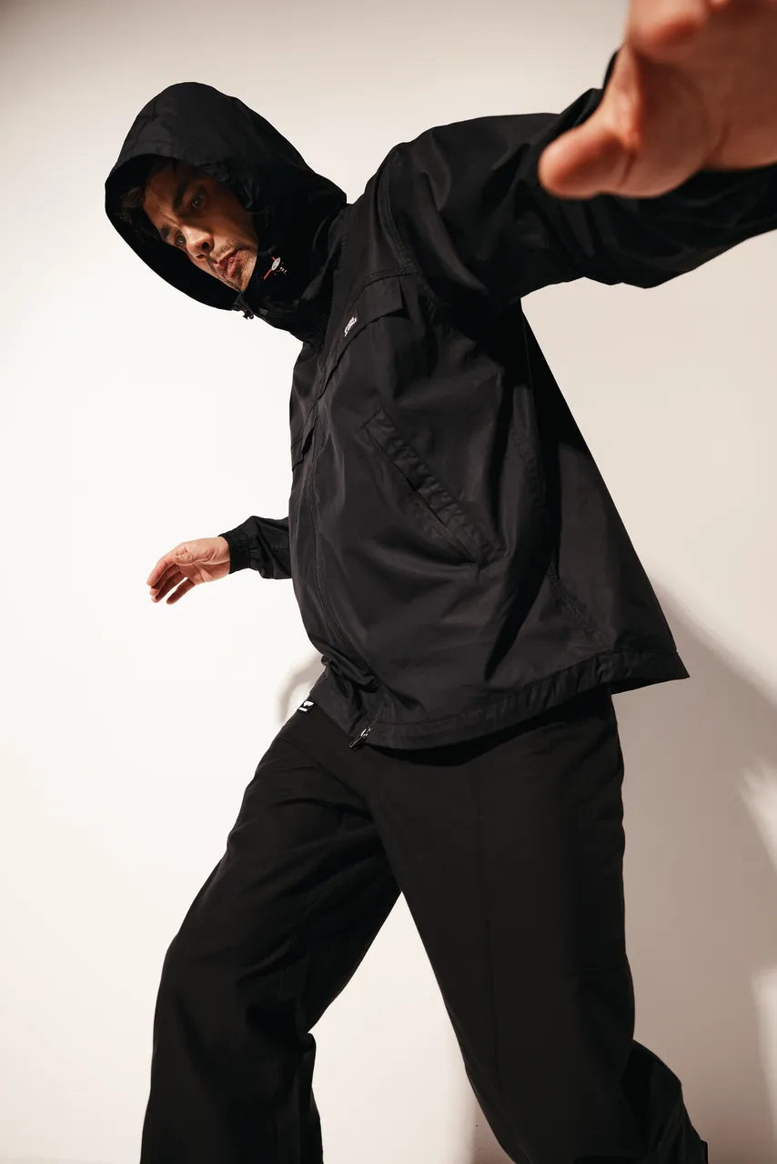 Model in a black hooded jacket and pants reaches toward the camera.