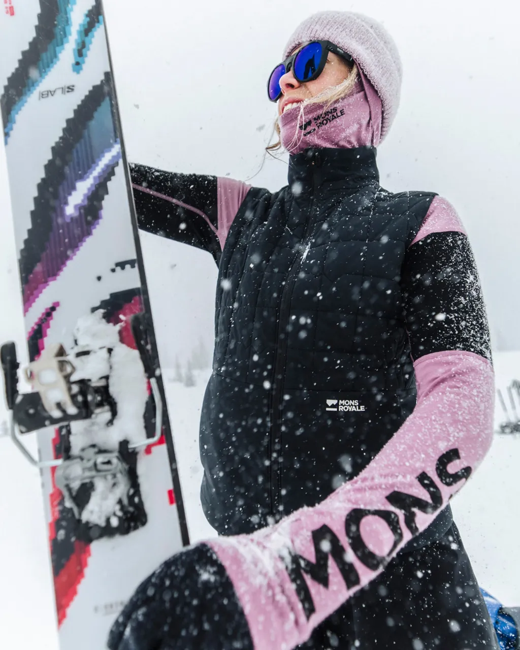 Skier in a black vest and pink gear holds a patterned ski as snow falls.