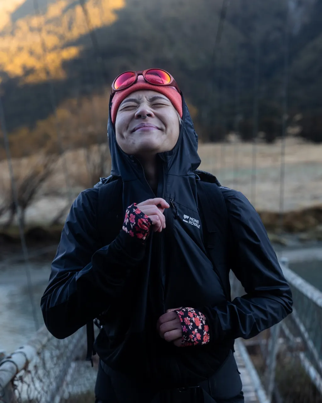Trail runner in a black hooded jacket and floral gloves smiles on a mountain path at sunrise.
