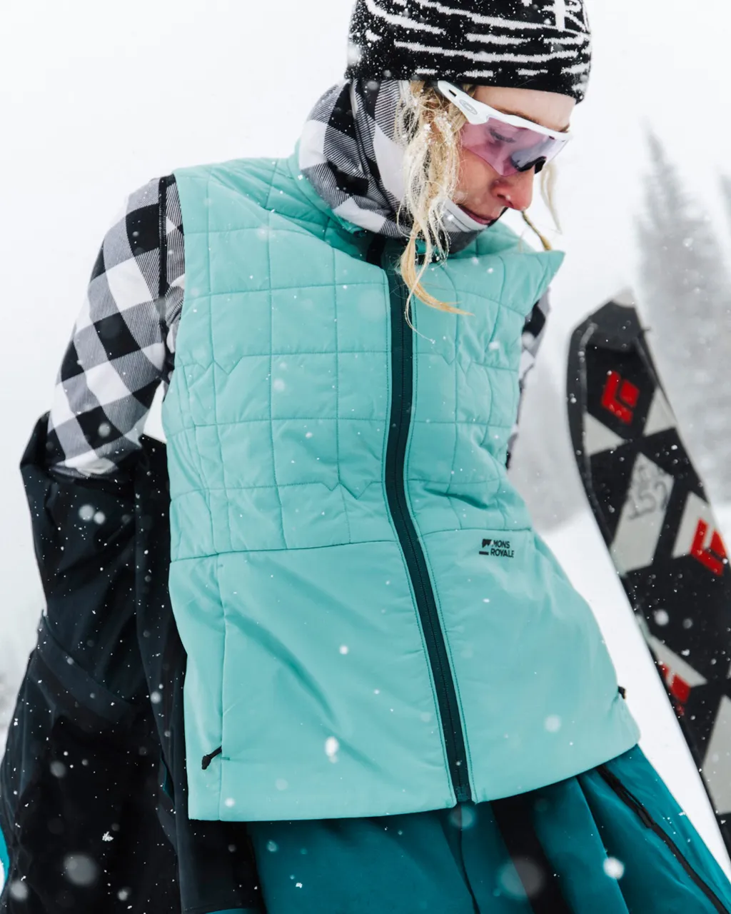 Snowboarder in a mint quilted vest and goggles stands in falling snow with a snowboard.