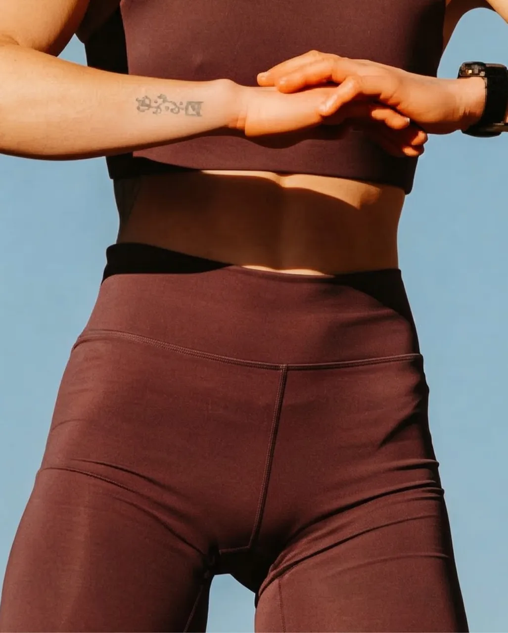 Athlete in a burgundy crop top and high-waisted leggings stretches her arms against a blue sky.