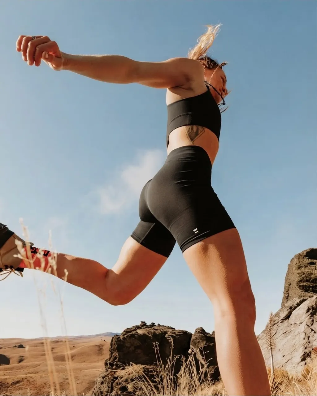 Athlete in black activewear stretches one leg on a rocky trail under a blue sky.
