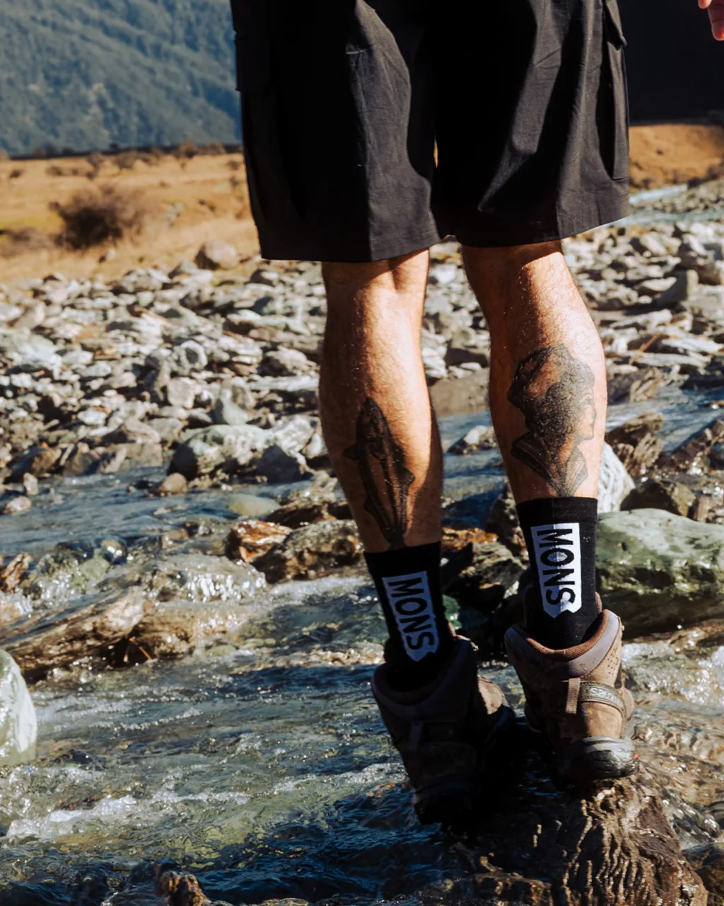 A person in Mons Royale socks and hiking sandals stands in a shallow rocky stream.