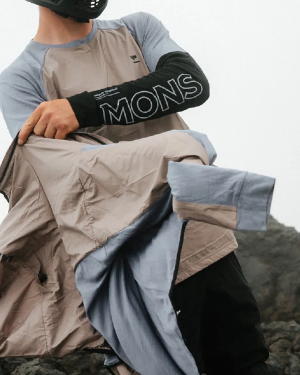 A mountain biker in a grey and taupe jersey takes off a lightweight jacket on a rocky hillside.