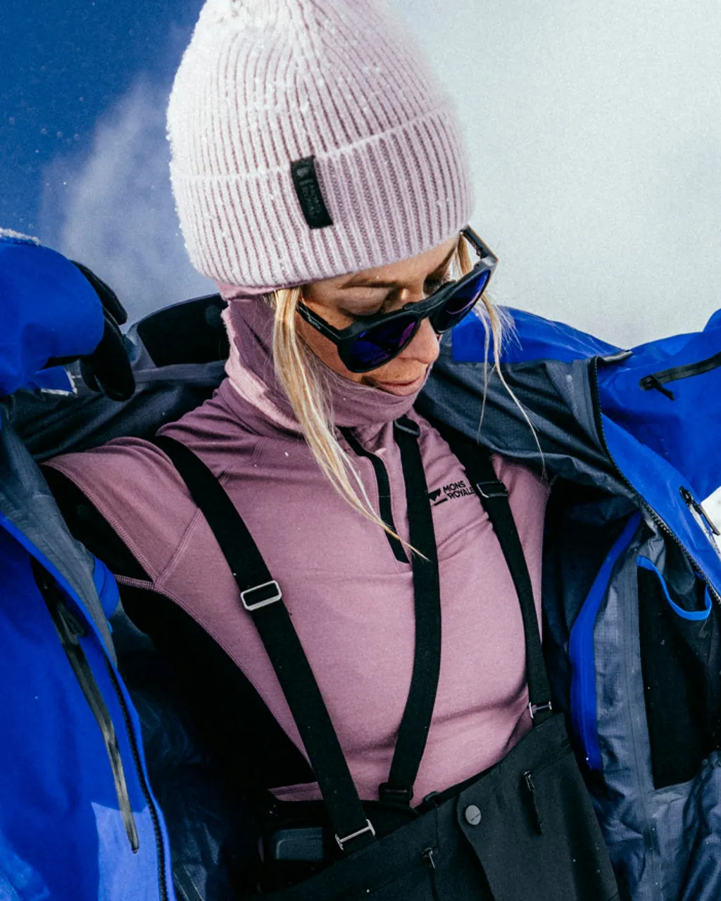 Woman in a pink beanie and base layer adjusts a blue ski jacket.