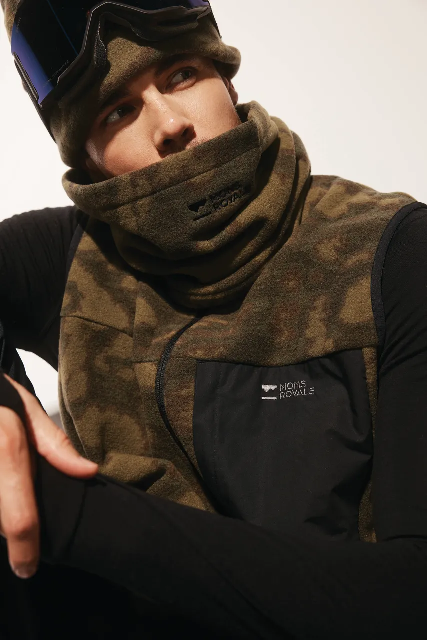 Model wears a brown camouflage fleece neck warmer and vest over black clothing.