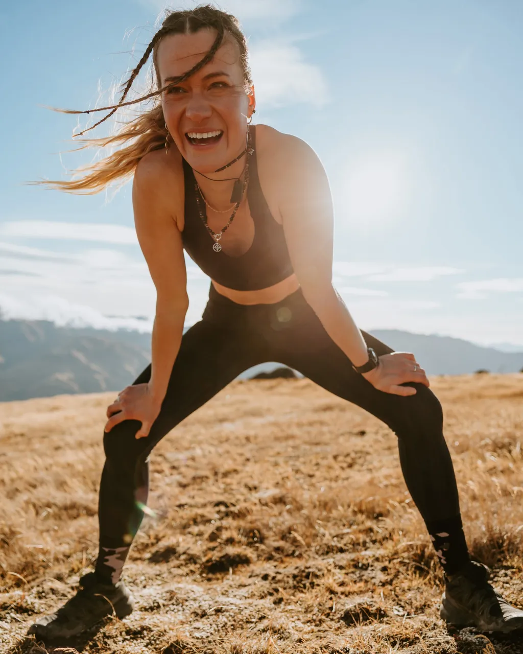 A smiling woman in black activewear rests on a sunny mountain trail.