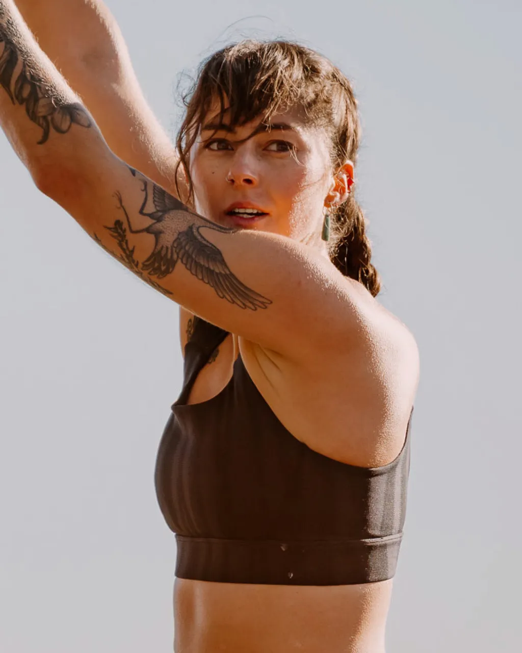 Tattooed woman in a dark brown sports bra raises her arms in warm sunlight.