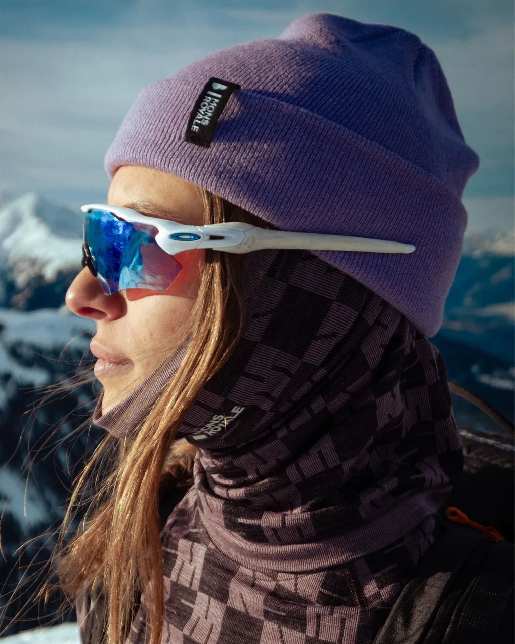 Woman in a purple beanie and mirrored sunglasses looks toward snowy mountains.