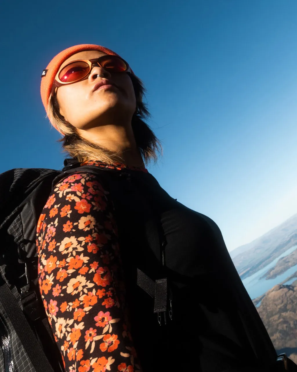 A woman in an orange beanie and floral top looks up at the sky, wearing a black backpack.