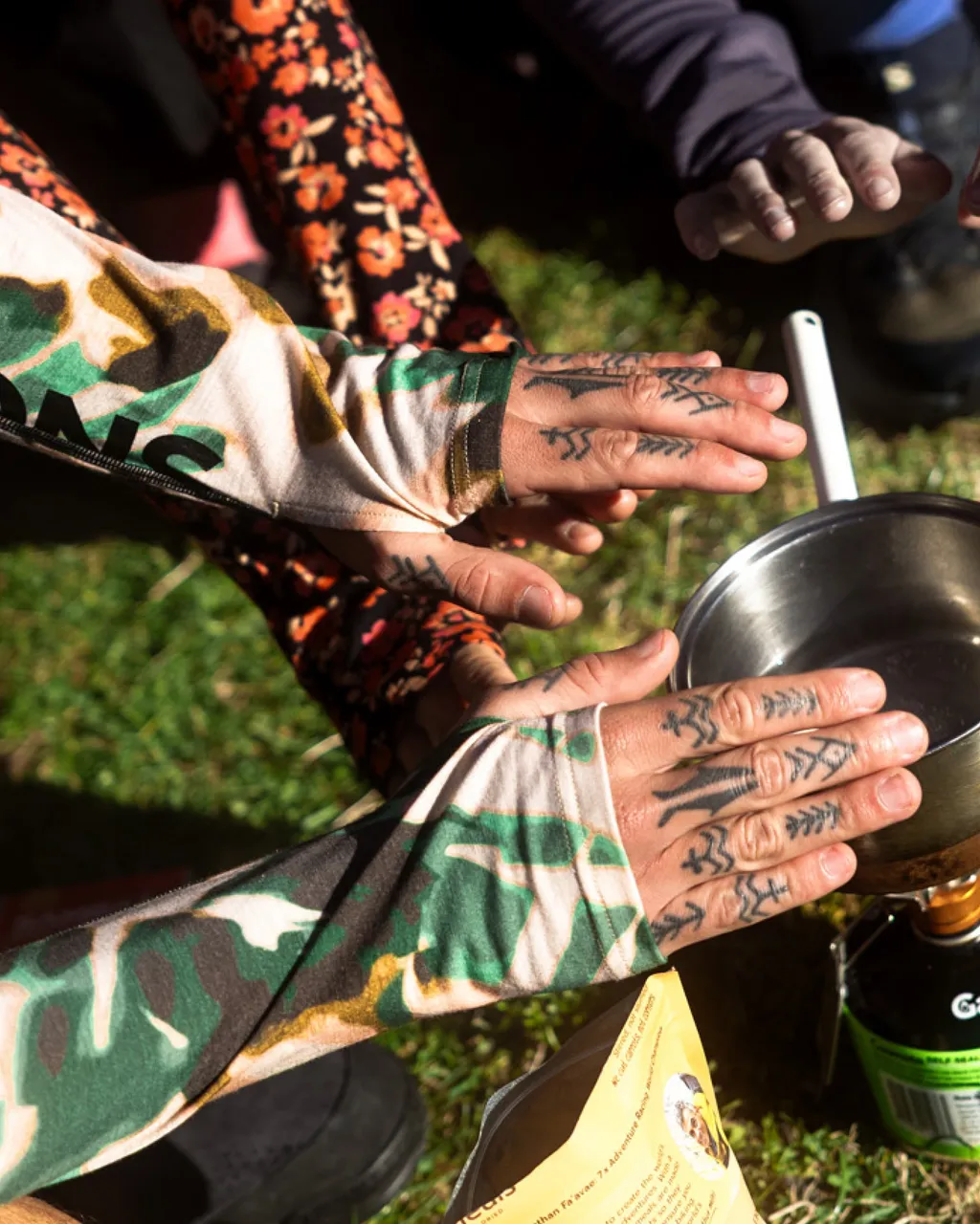 Tattooed hands in patterned sun sleeves gather around a metal camping pot outdoors.