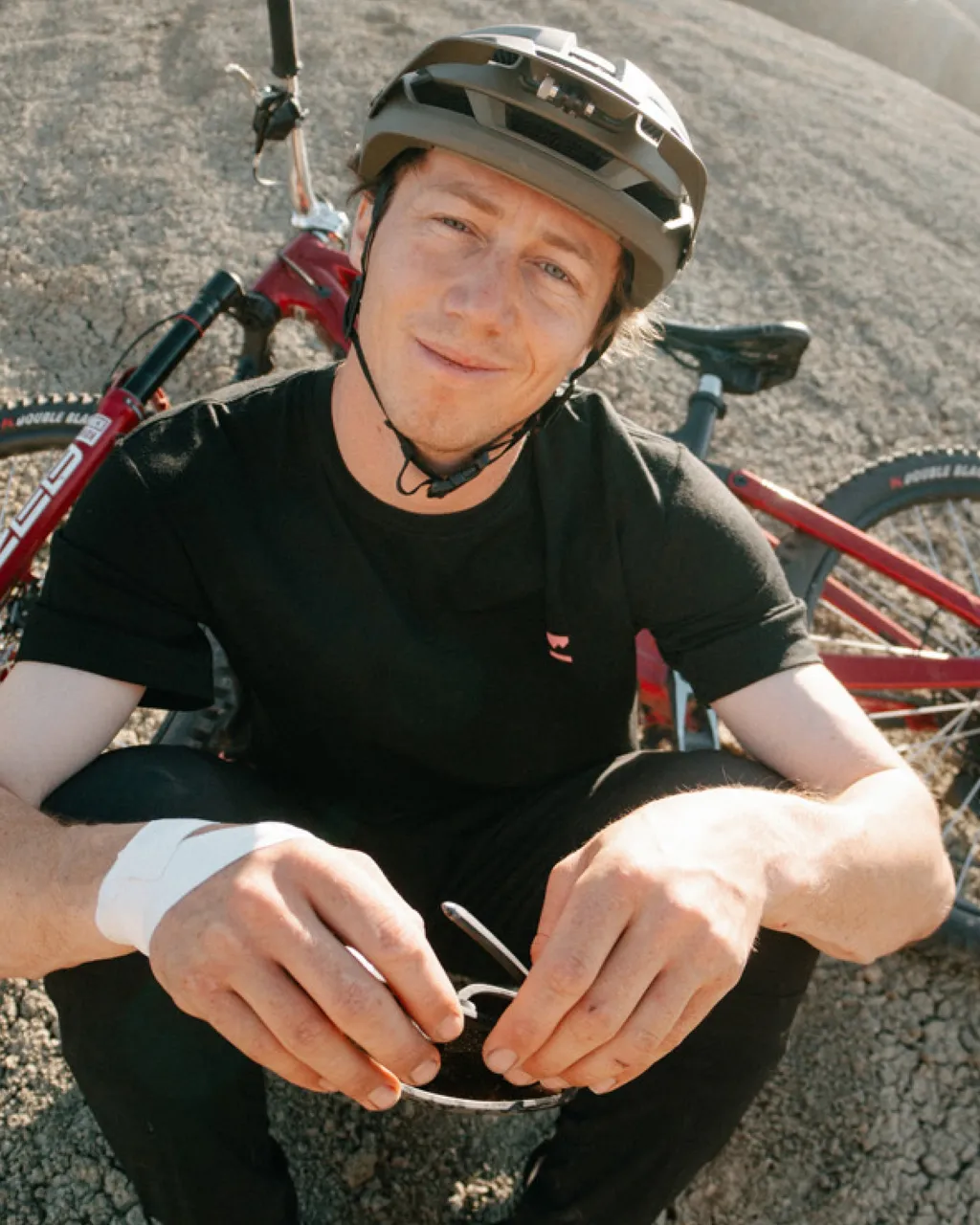 A helmeted mountain biker sits beside a red bike, holding sunglasses on rocky ground.