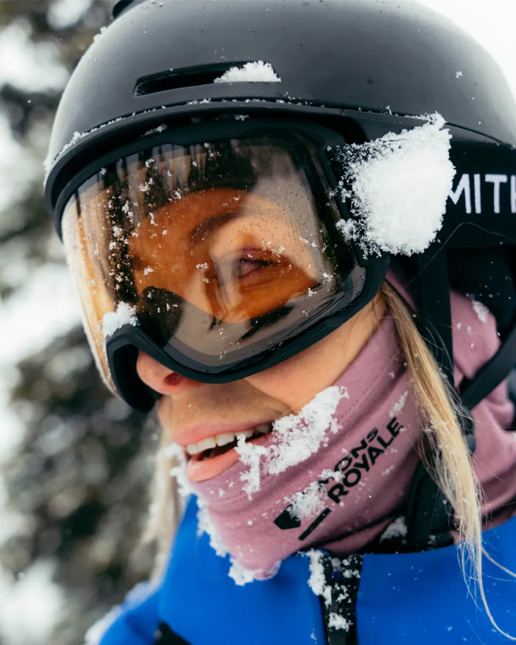 A skier in a black helmet and amber goggles smiles in falling snow, wearing a blue jacket and pink neck warmer.