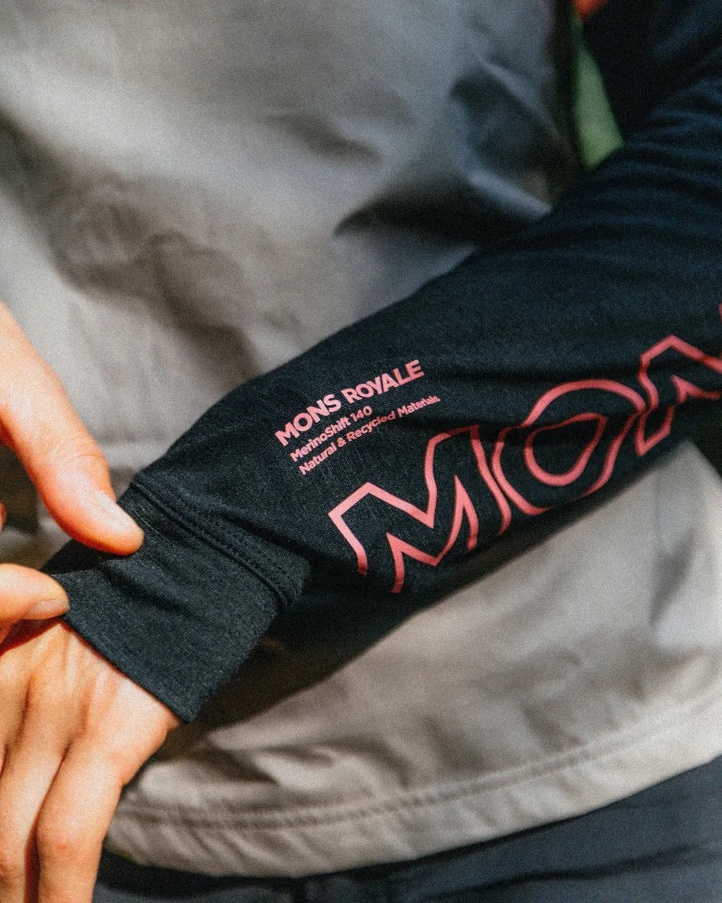 A hand holds out the black sleeve of a Mons Royale merino top, showing its pink logo and ribbed cuff.