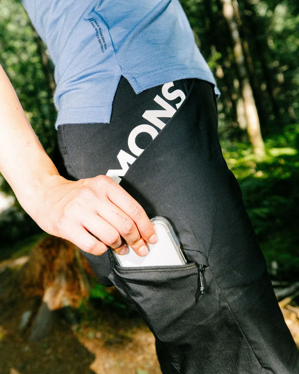 A person slides a phone into the zip pocket of black Mons Royale shorts outdoors.