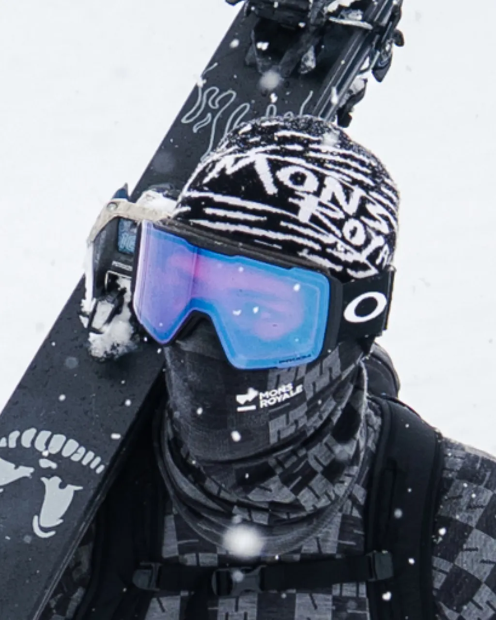 A skier in blue goggles and a patterned face covering carries a black ski over one shoulder as snow falls.