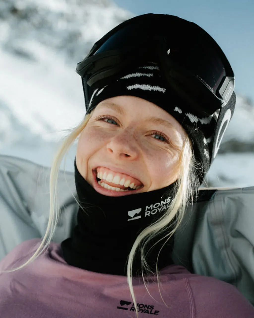 A smiling skier wears a black helmet, Mons Royale neck warmer and purple top against a snowy mountain backdrop.