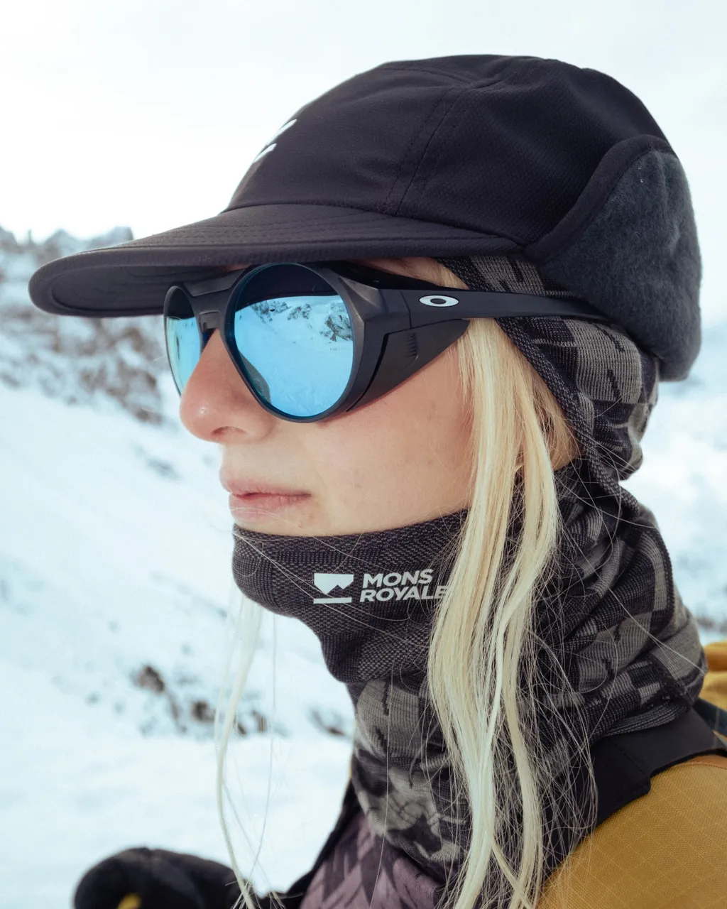 Woman in a black Mons Royale neck warmer, cap and blue mirrored goggles against a snowy landscape.