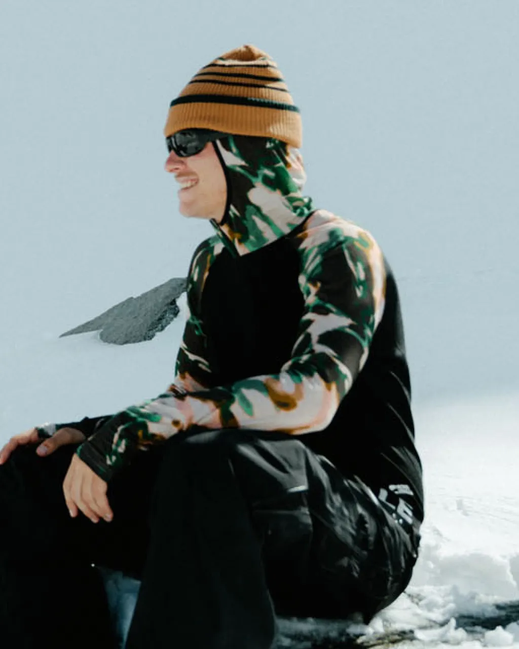 A seated snowboarder in camouflage gear and an orange striped beanie rests on the snow.