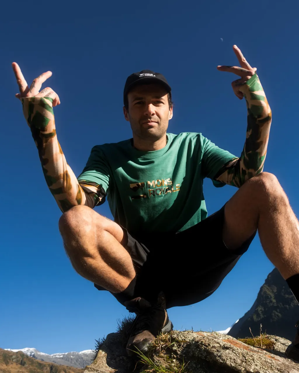 A man in a green Mons Royale T-shirt crouches on a rocky summit and raises both hands in peace signs.