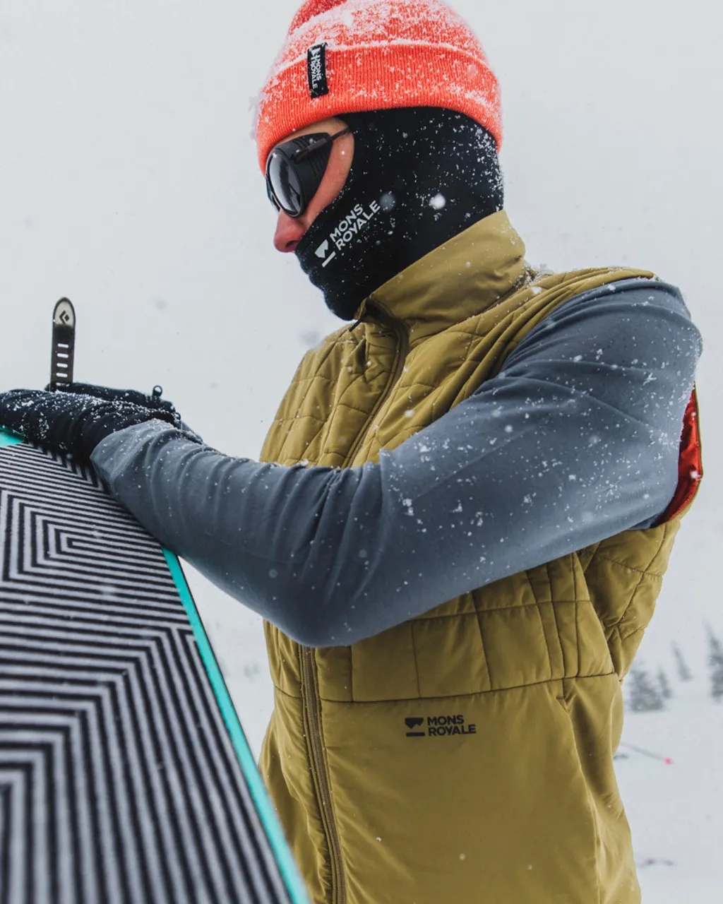 Snowboarder in a coral beanie and olive vest adjusts a snowboard in falling snow.