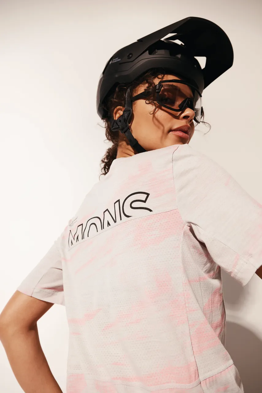 A woman in a black bike helmet and goggles wears a pale pink Mons Royale jersey.