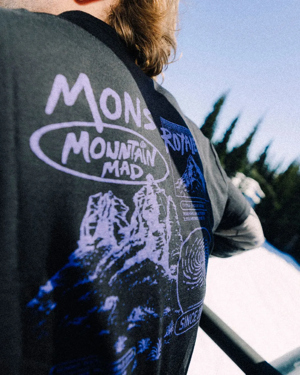 A person wears a charcoal Mons Royale T-shirt with purple mountain graphics.