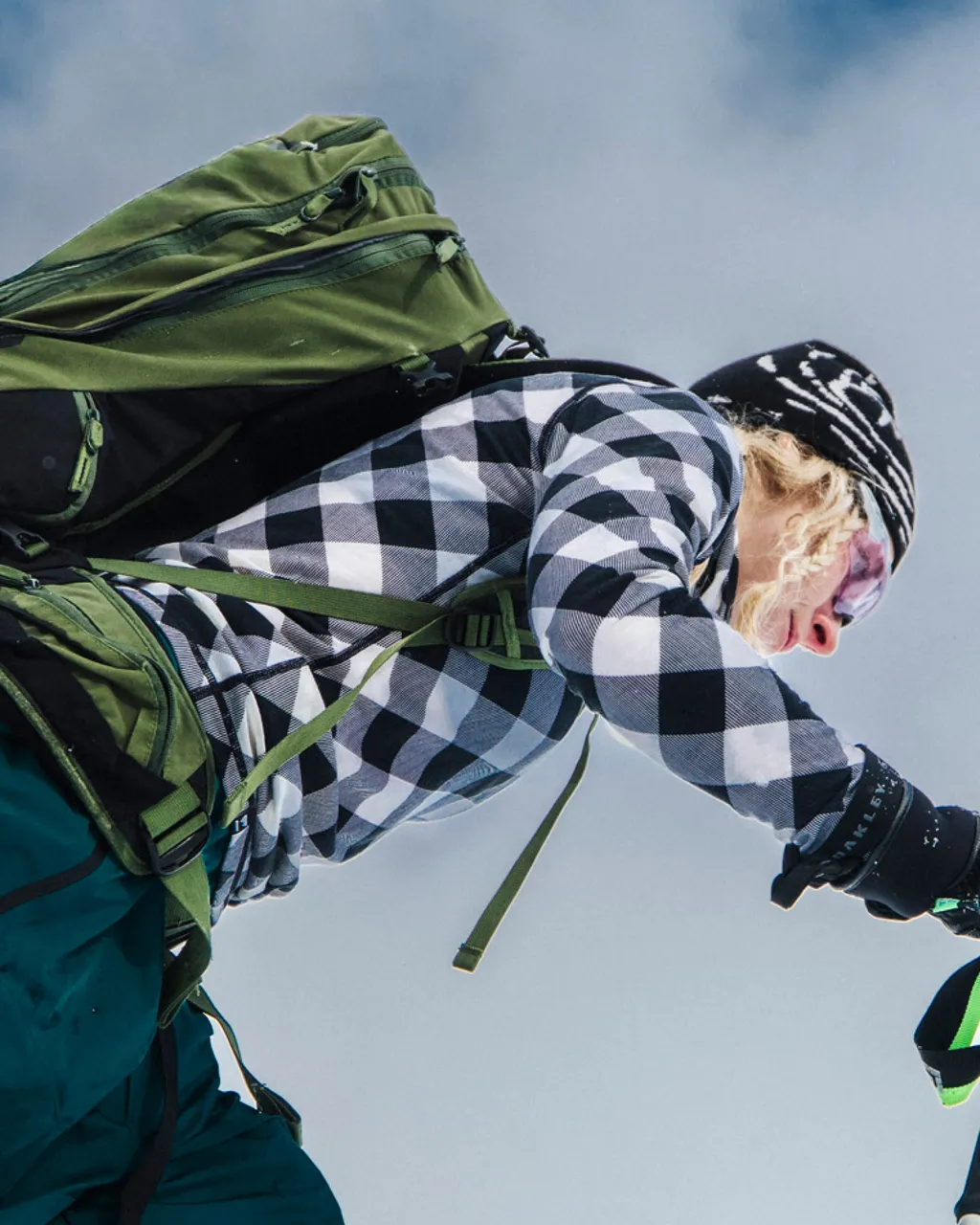 A skier wearing a checkered top and beanie climbs uphill with a large green backpack.