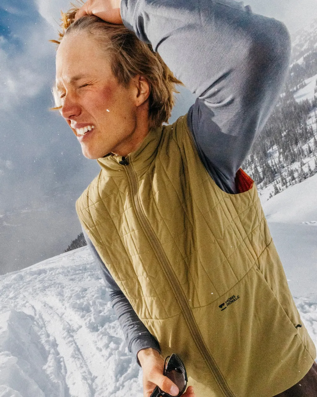 A man in a tan quilted vest stands in the snow with one hand resting on his head.