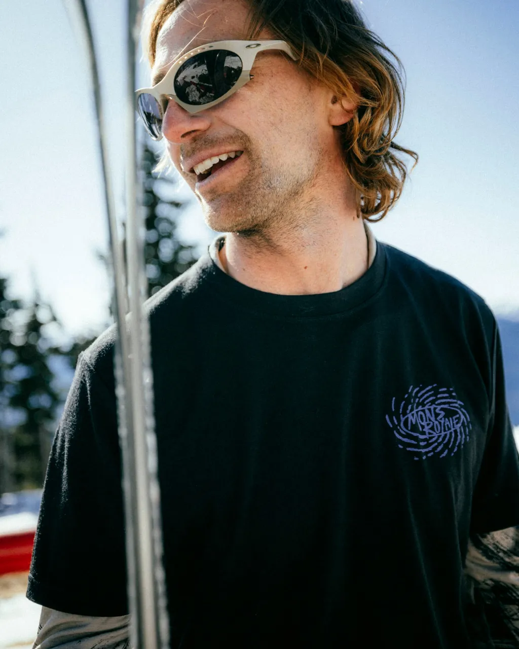 Man in white sunglasses smiles outdoors wearing a black Mons Royale T-shirt with a purple chest logo.