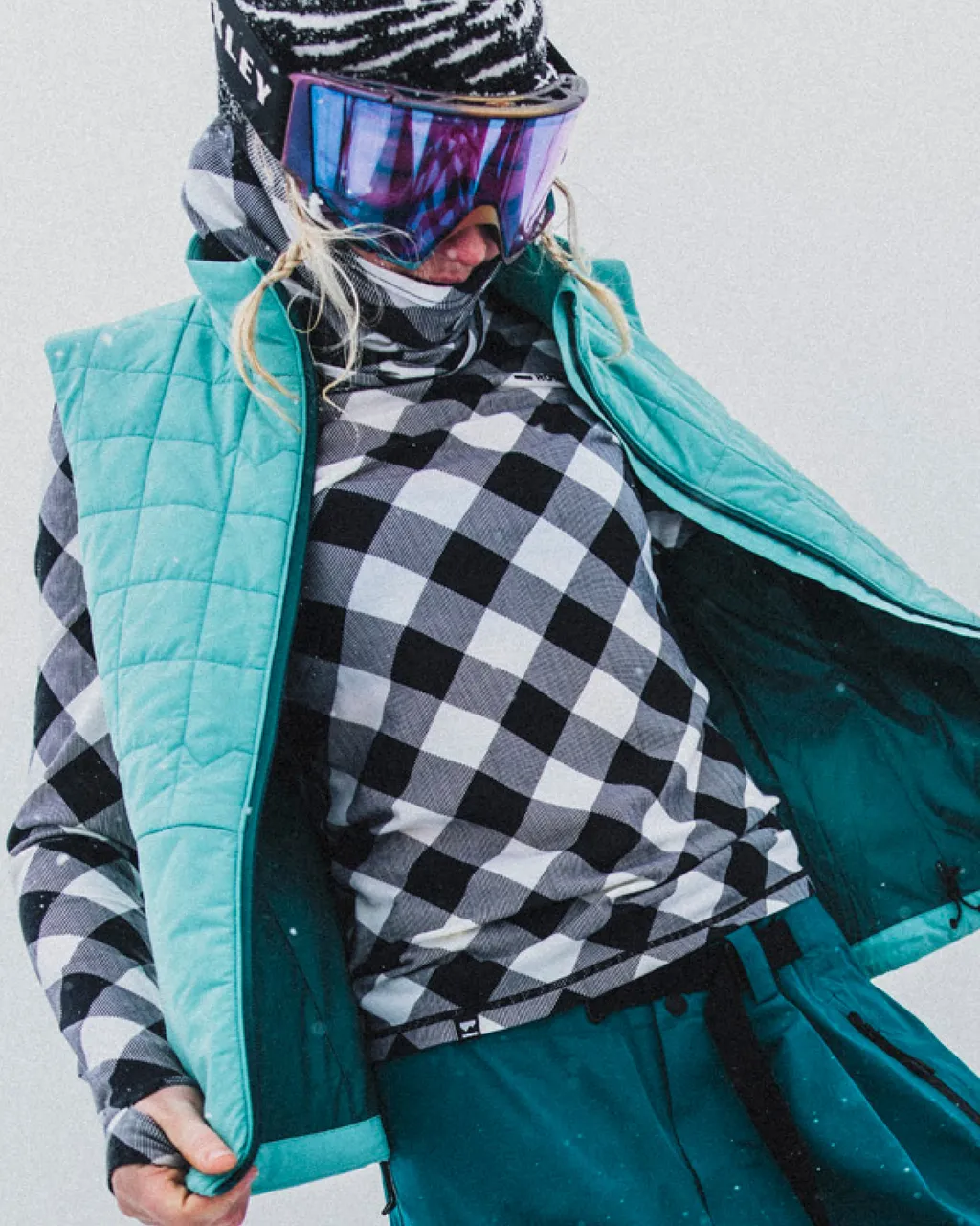 Snowboarder in a checkered base layer and turquoise vest adjusts the front of the vest.