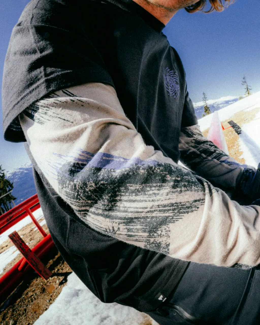 A person in layered graphic tops sits beside a red chair in a snowy mountain setting.
