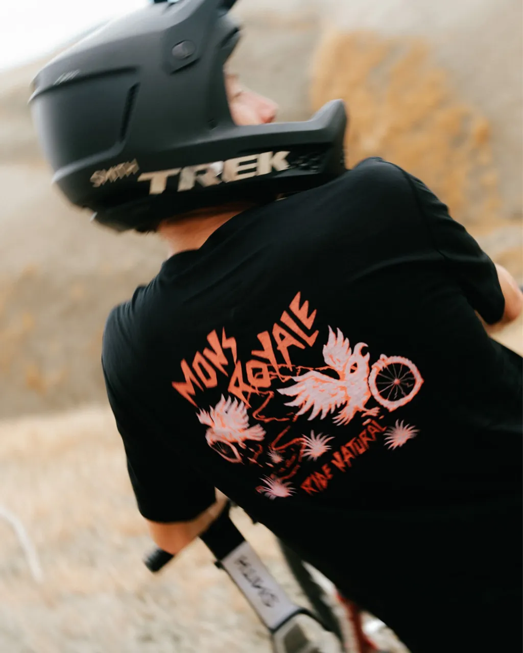 A mountain biker in a black Mons Royale graphic T-shirt and Trek helmet rides along a rocky trail.