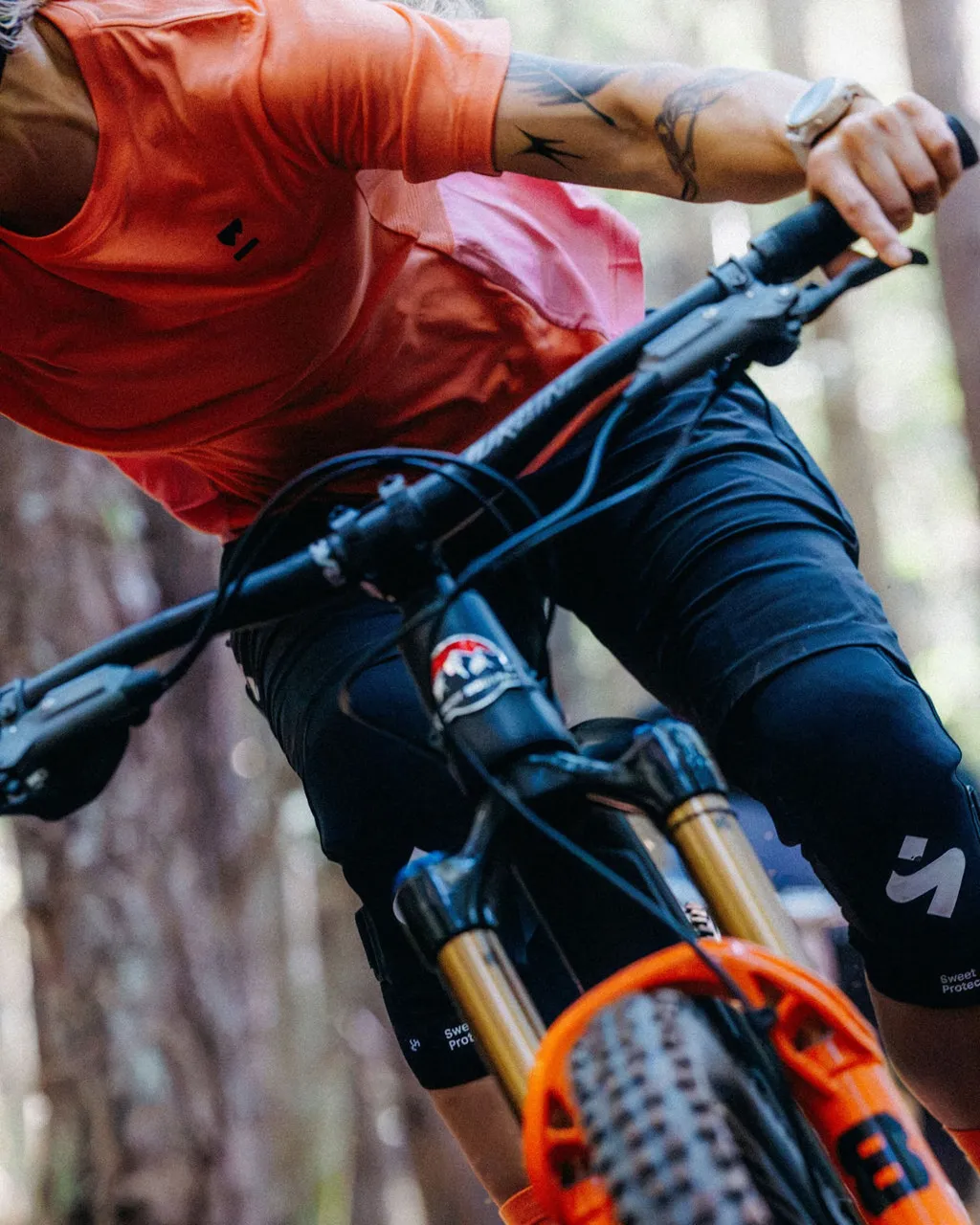 A mountain biker in an orange jersey rides through a wooded trail.