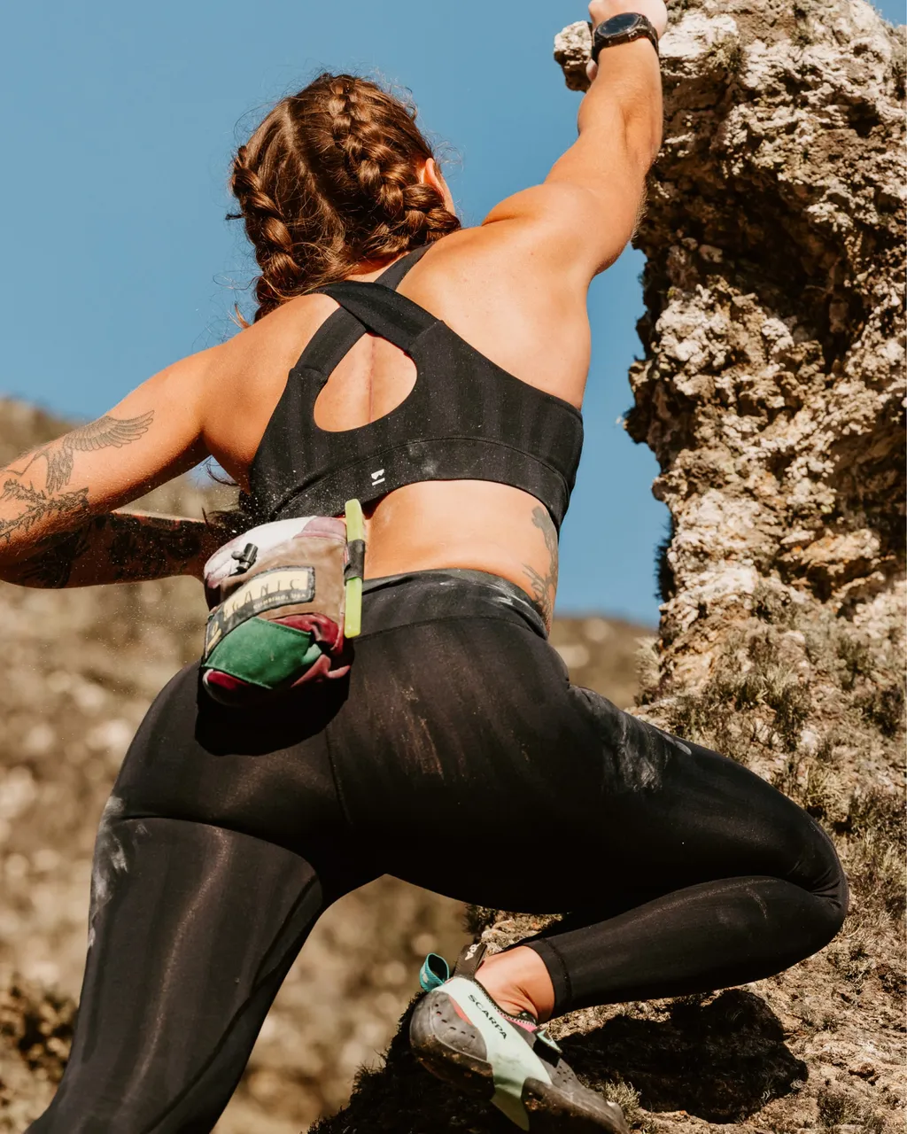 A woman in black activewear climbs a rocky face beneath a clear blue sky.