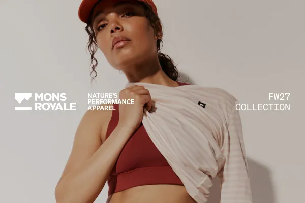 Model in a red cap and sports bra holds open a pale striped top for the Mons Royale FW27 collection.