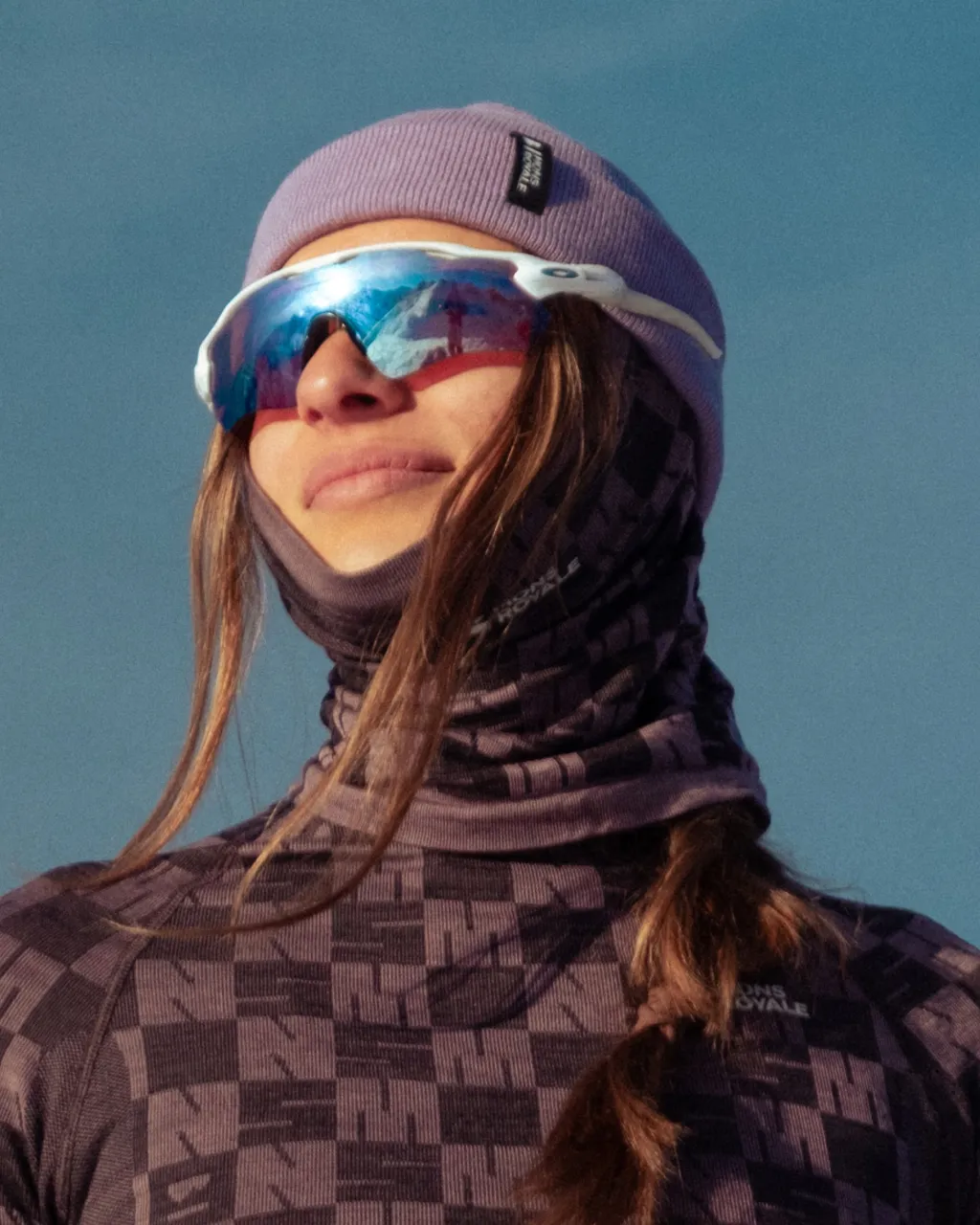 Woman in a purple beanie, mirrored ski goggles and patterned winter layers against a blue sky.