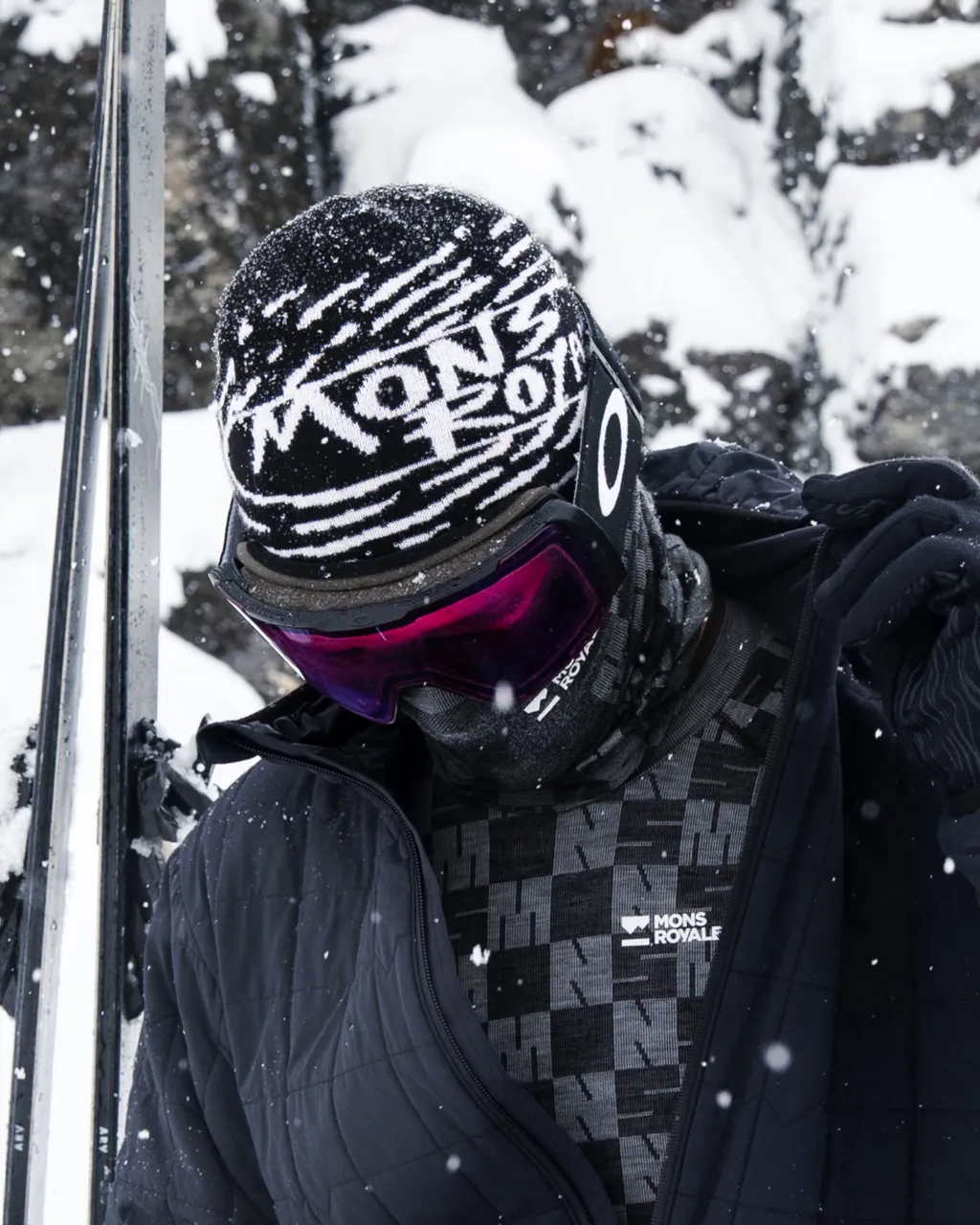 A skier in a Mons Royale base layer and patterned beanie adjusts their gear in falling snow.