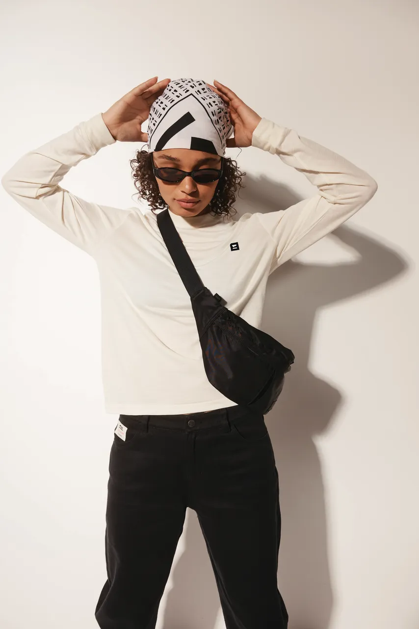 Model adjusts a patterned headscarf while wearing a white top, black trousers, sunglasses and a crossbody bag.
