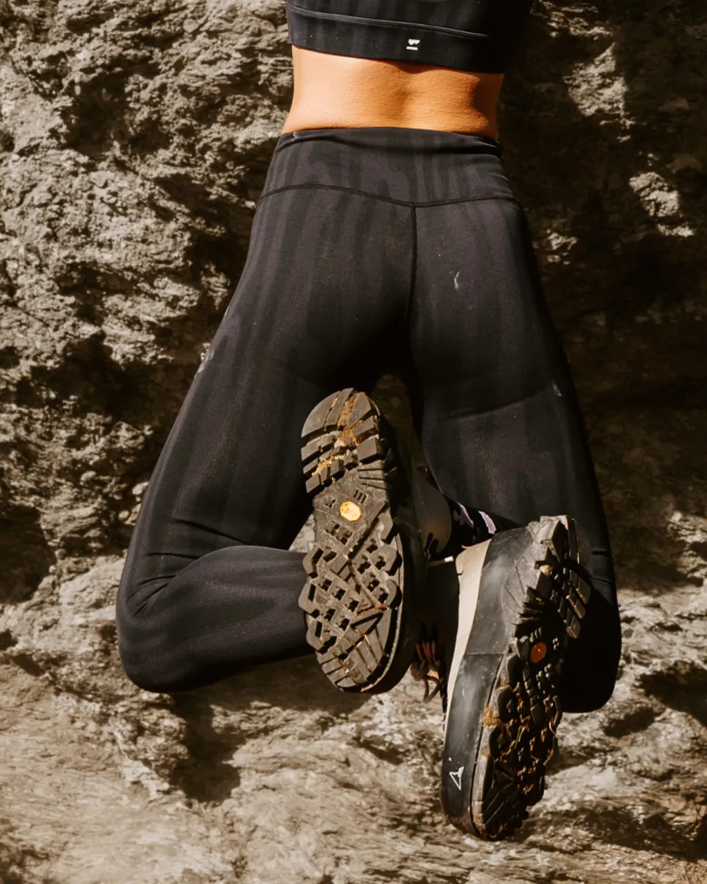 A hiker in black leggings kneels on rock, showing the tread on their trail shoes.