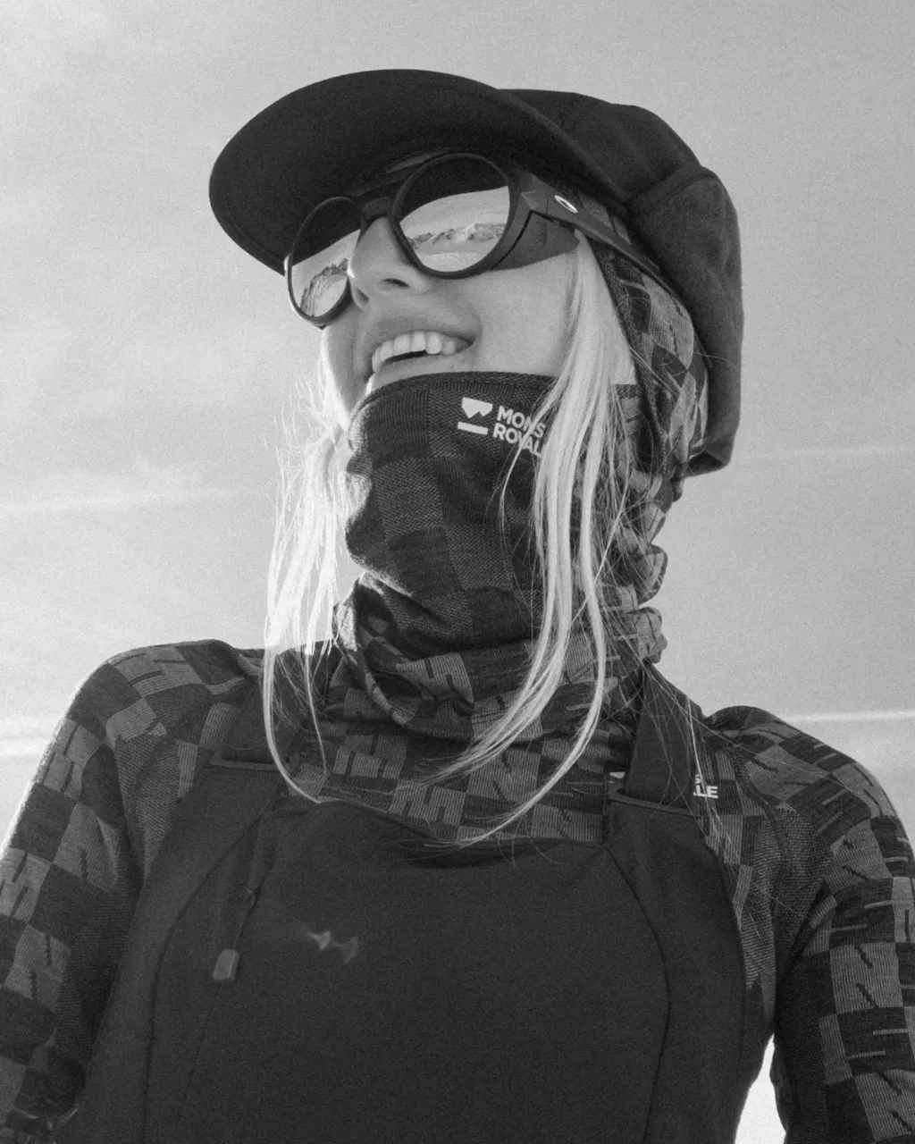 Smiling woman in a cap and mirrored sunglasses wears a Mons Royale neck gaiter and patterned outdoor clothing.