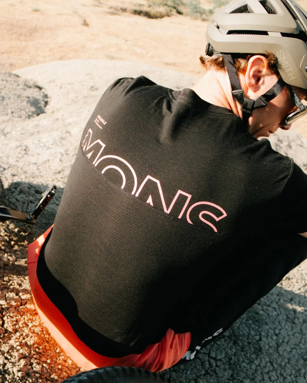 A helmeted cyclist in a black Mons Royale top crouches on rocky ground beside a bike.