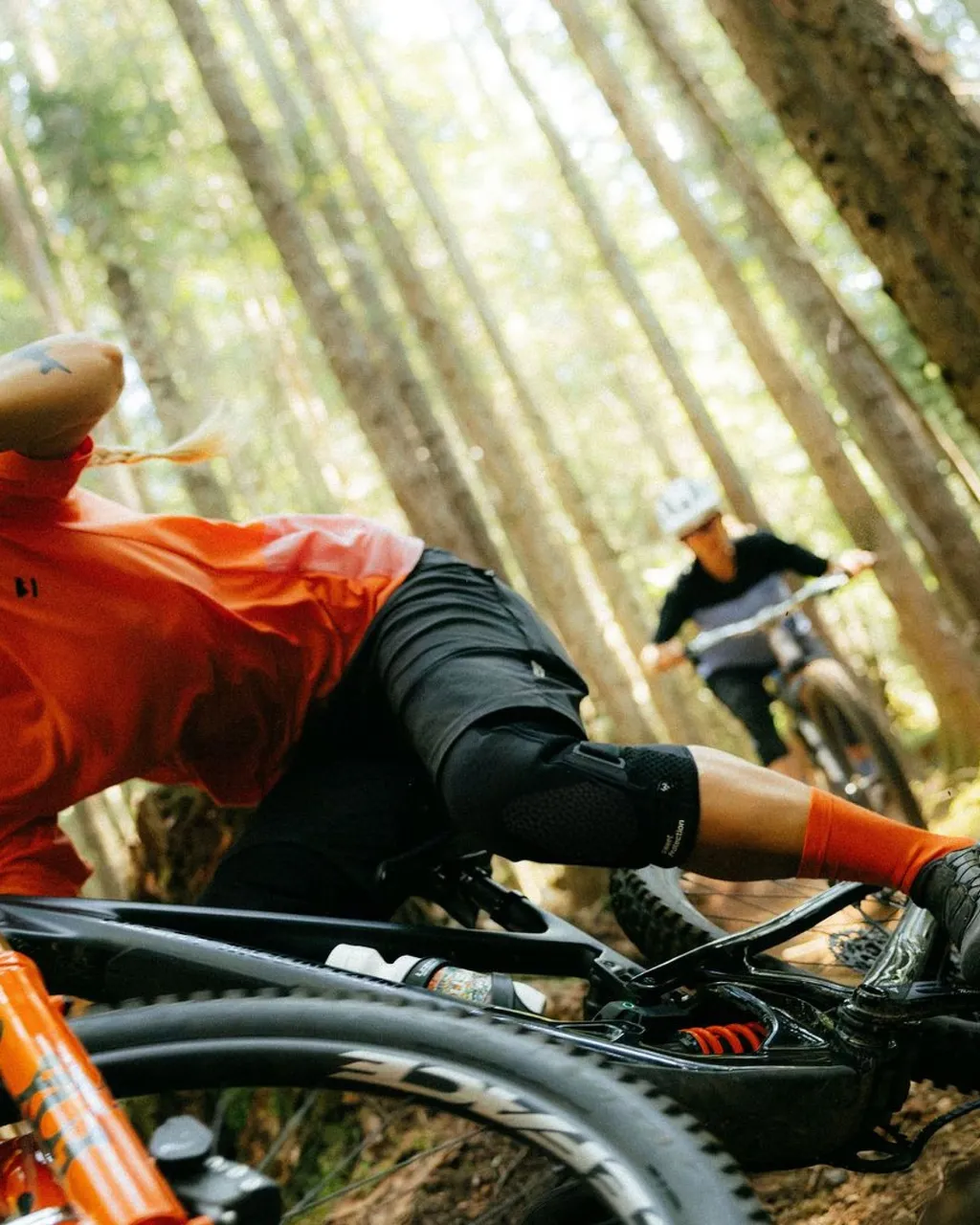 A mountain biker in an orange jersey leans over a bike on a wooded trail.