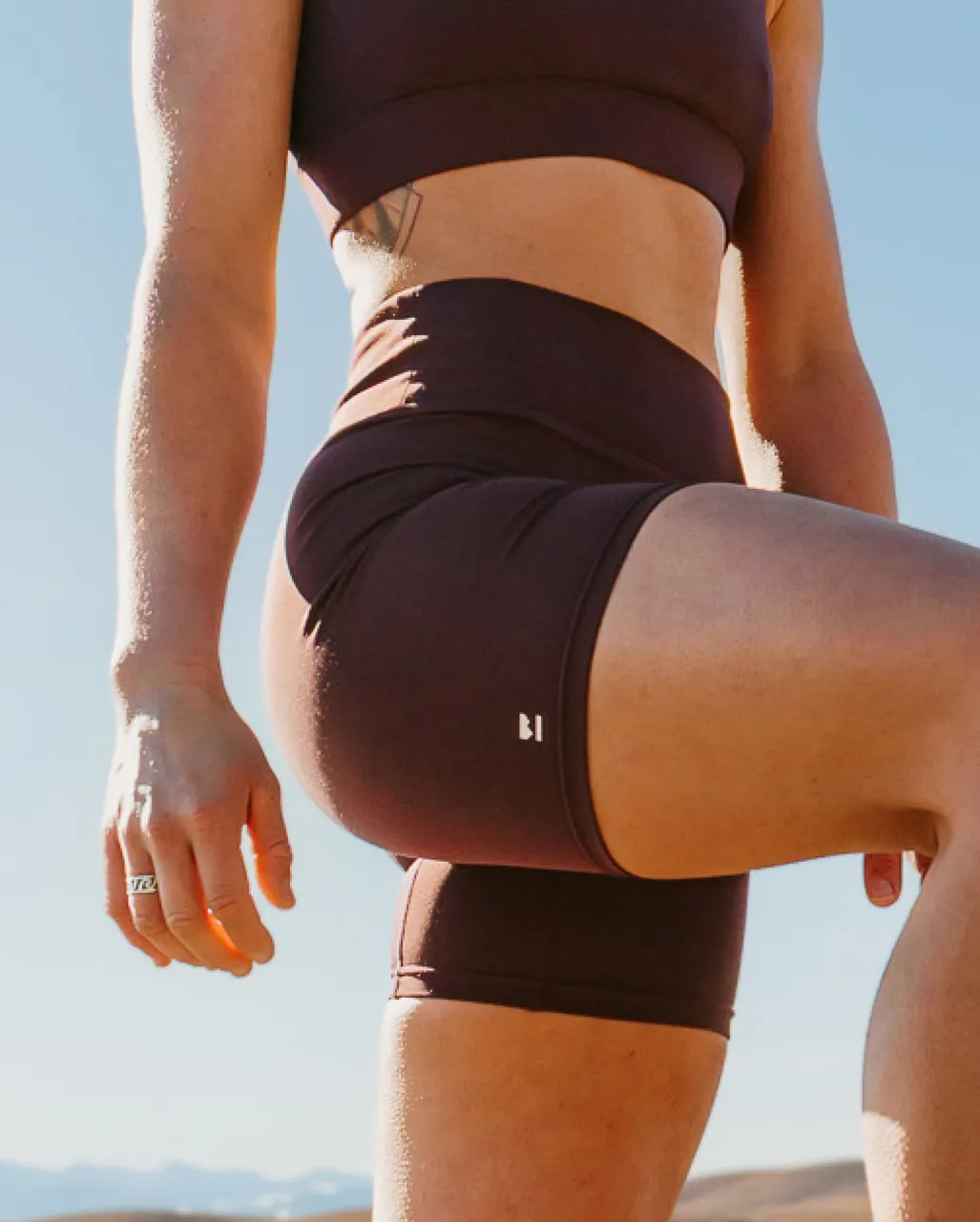 Athlete in dark brown sportswear raises one knee outdoors in bright sunlight.