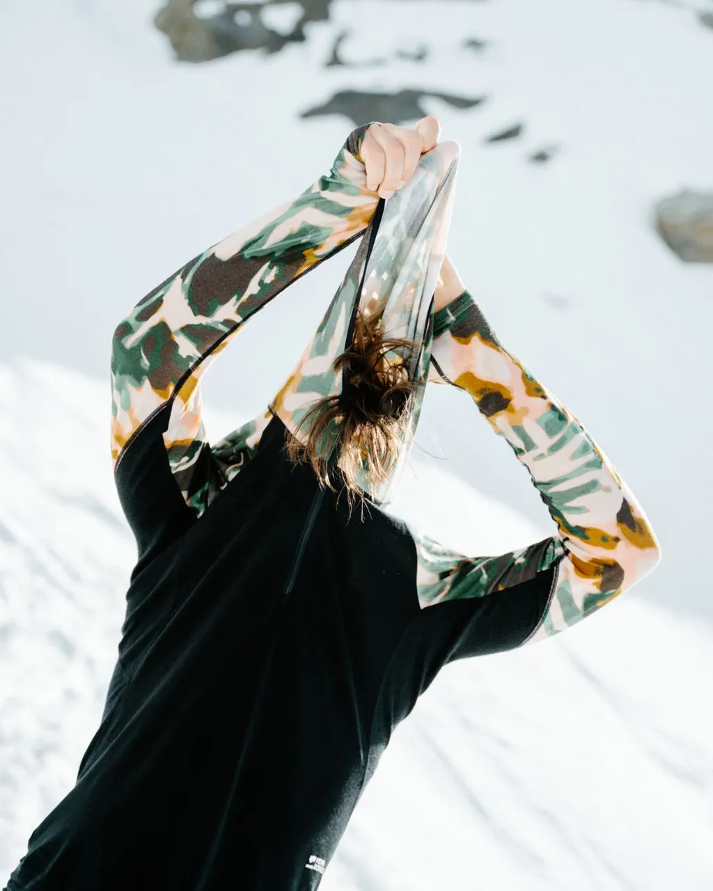 Woman in a black top with camouflage sleeves ties her hair in the snow.