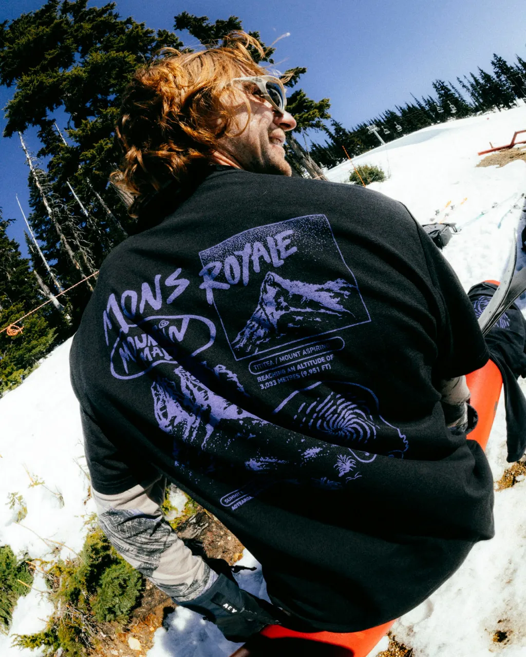 A skier in sunglasses wears a black Mons Royale graphic T-shirt on a snowy slope.