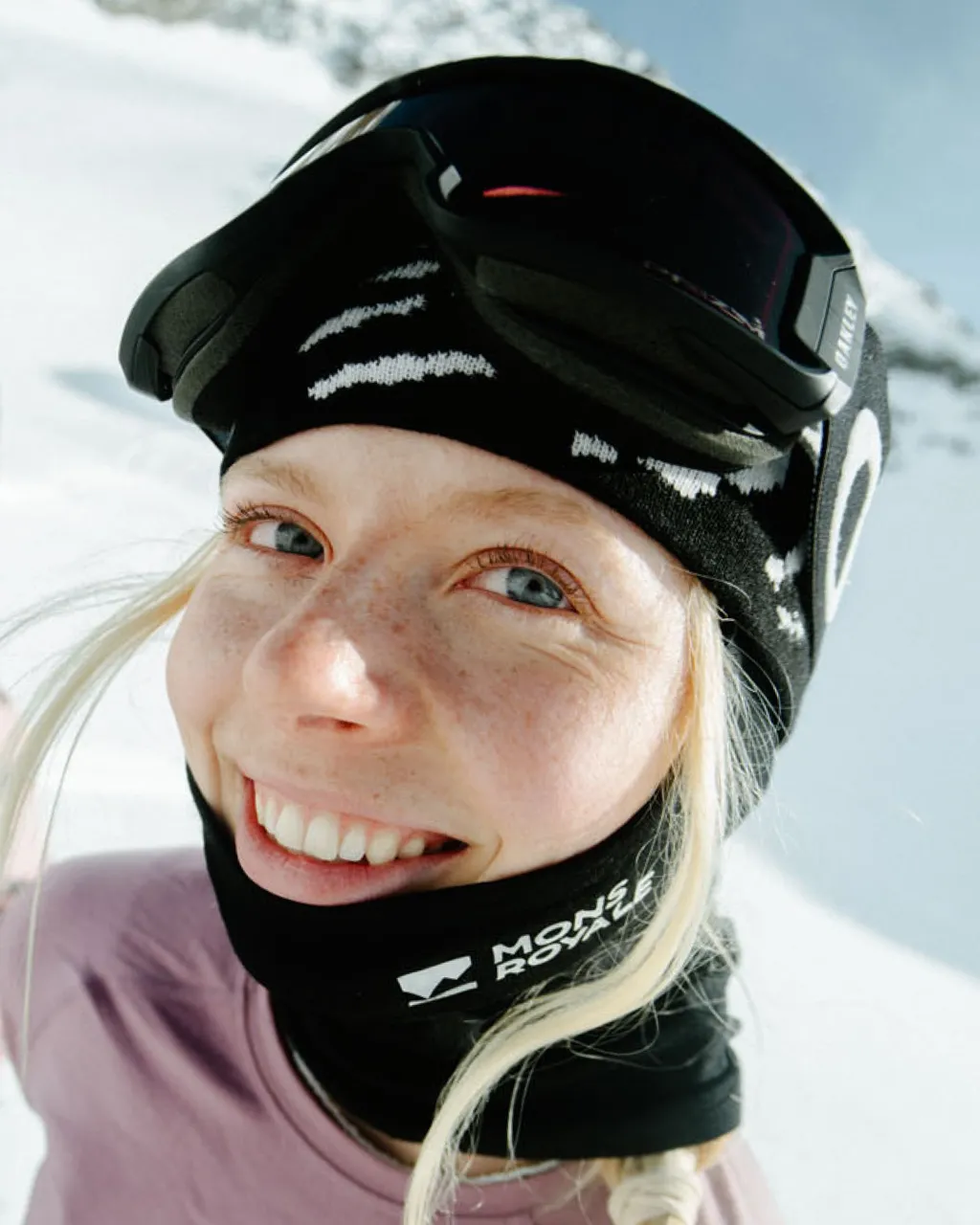 Smiling skier wears black Mons Royale neckwear and a patterned headband with goggles pushed up.