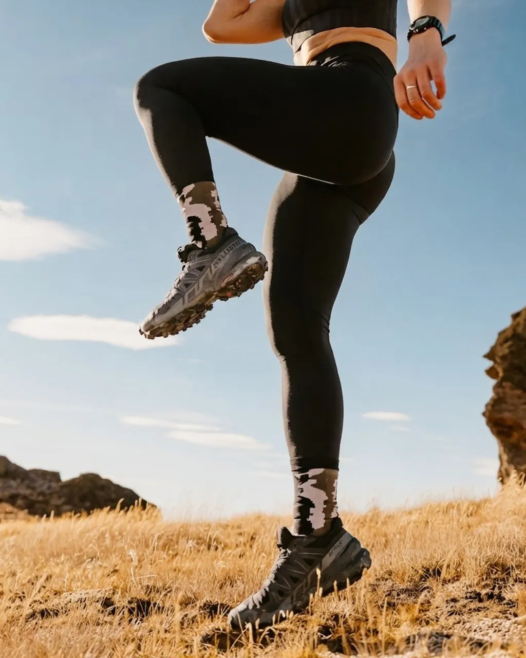 Trail runner in black leggings and patterned socks strides across a golden grassy hillside.