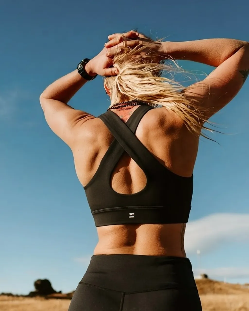 A woman in a black cross-back sports bra stretches with her hands above her head outdoors.