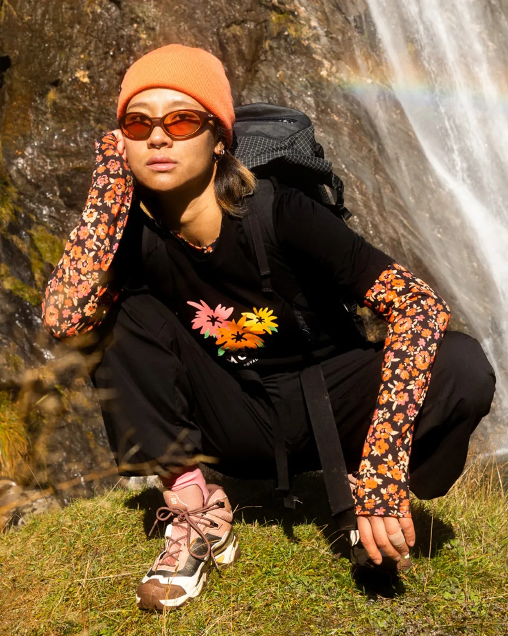 Hiker in an orange beanie crouches beside a waterfall wearing floral arm warmers and a backpack.